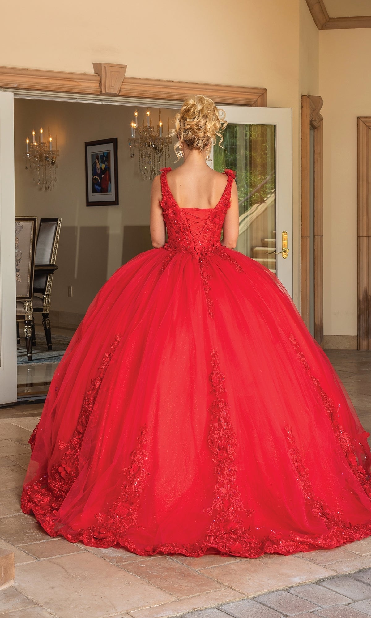 A woman with blonde hair in an updo wears the Quinceanera Dress 1678 By Dancing Queen, a red, floor-length ball gown with a full skirt and embroidered details, standing indoors and facing away from the camera.